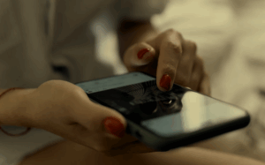 A person is holding a smartphone, scrolling through a social media feed. Their fingers are poised over the screen, with a focus on the device and hand.