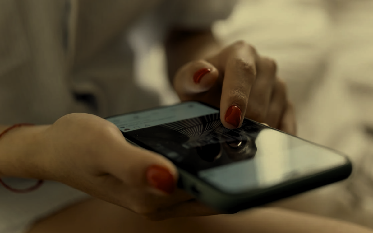 A person is holding a smartphone, scrolling through a social media feed. Their fingers are poised over the screen, with a focus on the device and hand.