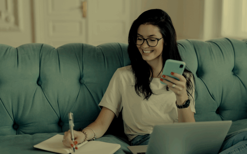 A woman sitting on a couch, smiling while using a smartphone and laptop, suggesting a balanced approach to digital connectivity.