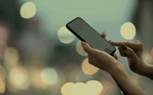 A person's hands using a mobile phone against a backdrop of soft, defocused lights, highlighting the integration of technology in leisure or social contexts.