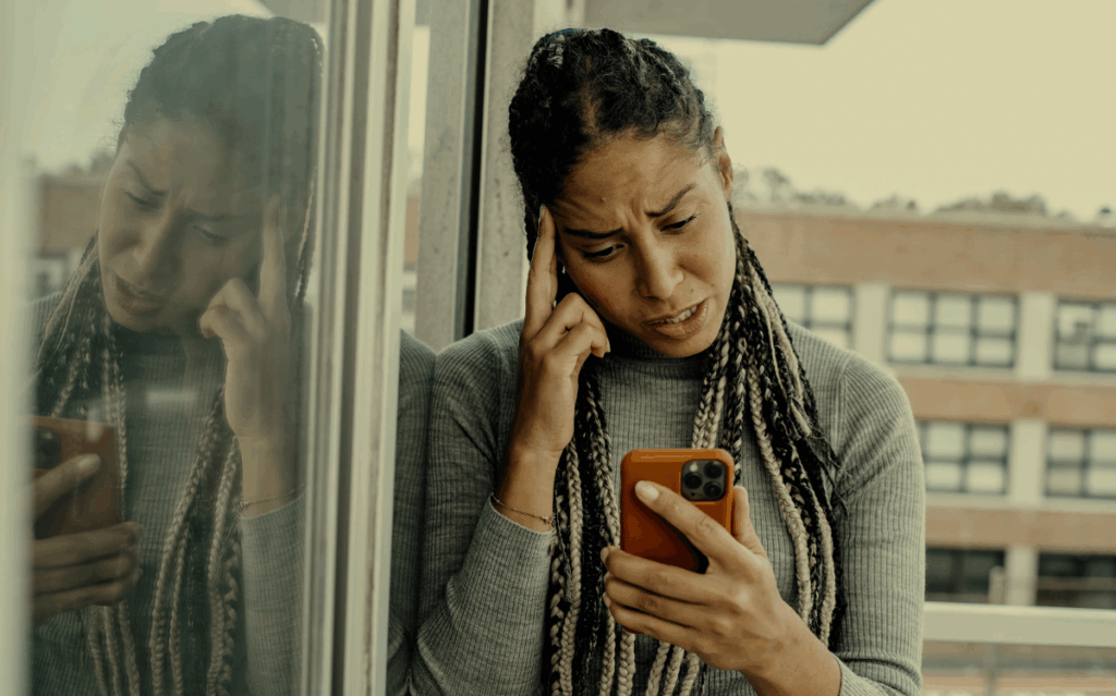 A person with braided hair appearing distressed while using a mobile device, suggesting the potential for technology to create emotional challenges.
