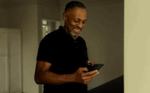 A man with a beard smiling while using a mobile device, indicating the role of technology in enabling communication and connection.