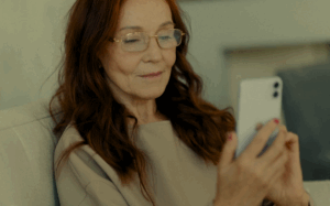 The woman is holding a smartphone in her hands, with her eyes slightly closed and a peaceful expression on her face. She is dressed in a beige top and is seated in a cozy indoor setting.