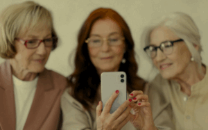A group of three women, two with white hair and one with red hair, are posing for a photo, smiling warmly. The focus is on the woman in the center holding the phone, with the others leaning in close.